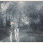 A ghostly black and white image of bare trees along. road with a ghostly superimposed image of a ghost and jack 'o' lantern.
